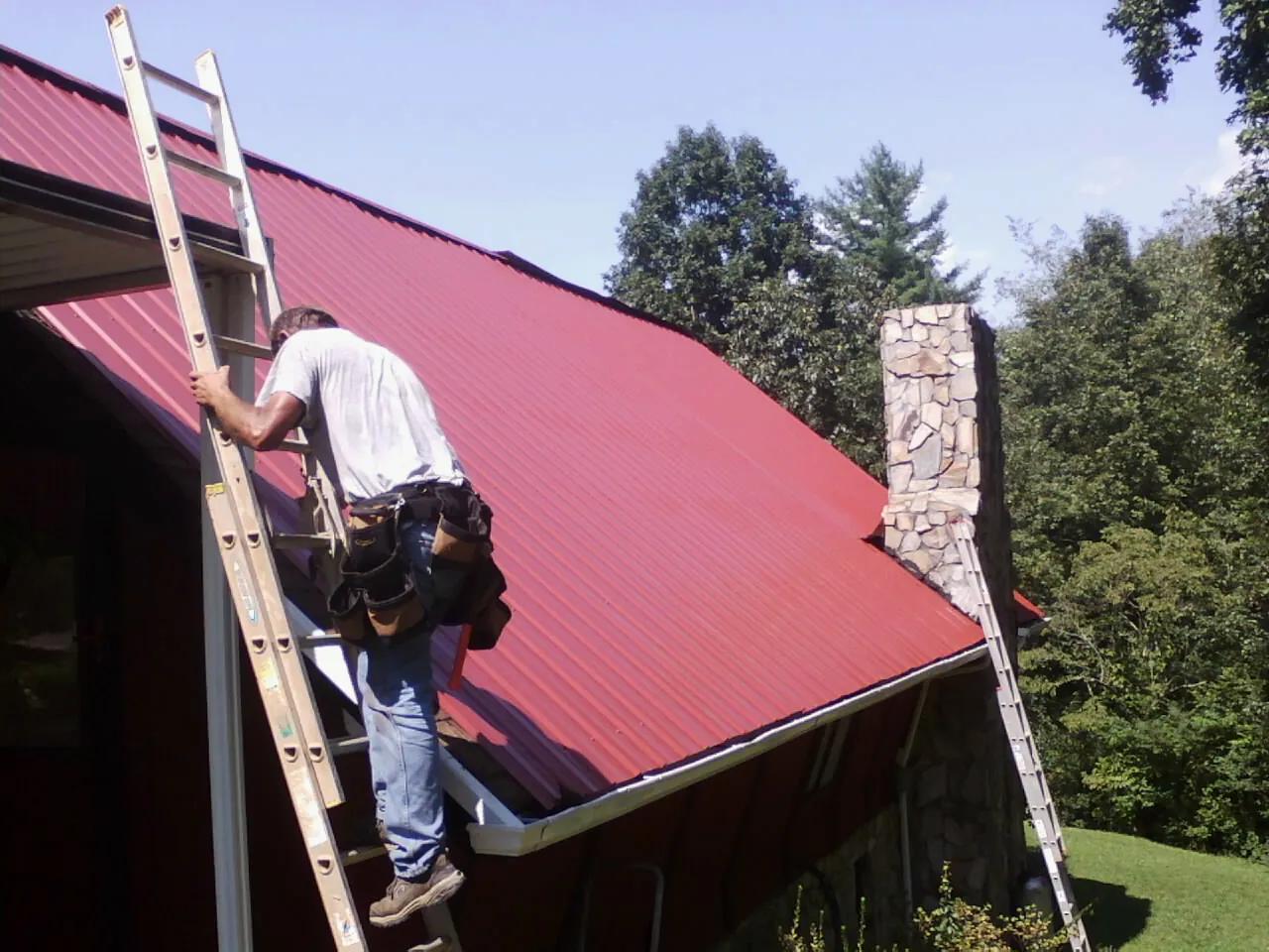 Roofer on a ladder beside a red metal roof and stone chimney — Soffit & Fascia Repair by Roofing Brothers in West Middlesex
